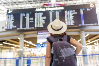 A woman wearing a straw hat and a backpack is standing in front of a large airport display. She is
