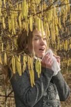 Young Caucasian woman with allergy problem stands by flowering hazel bush and sneezes into a