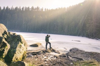 Person standing on the shore of a frozen lake surrounded by forest and sunbeams, Glaswaldsee, Bad