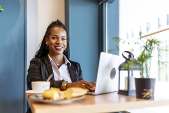 Latin casual businesswoman smiling at camera while using laptop having breakfast in a cafeteria