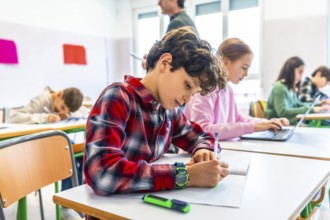 Elementary school students are diligently working at their desks, some writing in notebooks and