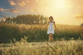 Happy young woman walking along country road in the meadow during sunset