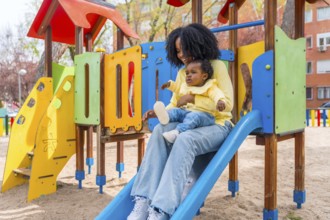 Happy African mother with baby girl on lap sliding in a colorful playground