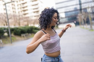 Curly haired hispanic woman joyfully dancing in an urban setting, embracing the sunny day with