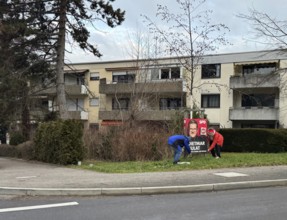 A scene in a residential area with trees, pavement and people working on an election poster,