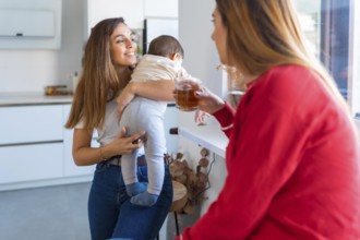Mother holding her baby while talking with a friend drinking tea in a modern kitchen, enjoying a