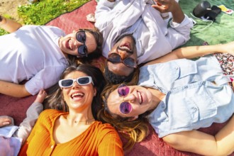 Four cheerful young adults lying together on blanket, enjoying sunny day picnic in park, listening