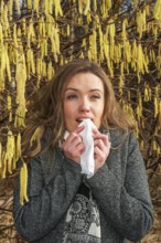 Young Caucasian woman with allergy problem stands by flowering hazel bush and sneezes into a