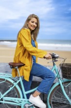 Young woman with yellow raincoat sitting on a turquoise vintage bicycle by the beach, enjoying a
