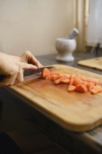 A person skillfully slices fresh carrots into bite-sized pieces on a wooden cutting board. Sunlight