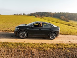 A driver sits in a black car on a dirt track, surrounded by green meadows, electric car VW ID5,