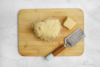 Shredded cheese on a wooden cutting board alongside a block of cheese and a grater. The items are