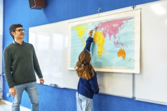 Smiling teacher watching elementary school student pointing at a world map during geography class