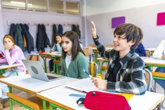 Smart young boy raising hand in classroom to answer question during lesson while classmates using