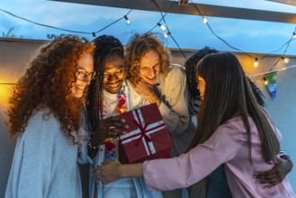African man opening birthday present embraced by his multiracial friends during rooftop party