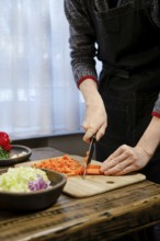 Unrecognizable man is chopping vibrant carrots on a wooden cutting board while surrounded by fresh