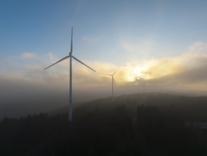 Wind turbines in the fog with the sun rising behind the trees, Rems Valley, Baden-Württemberg,