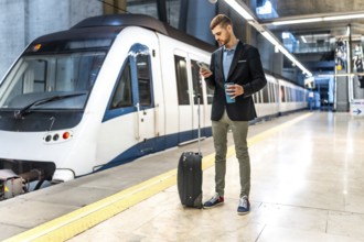 Young businessman standing on subway platform with suitcase, using smartphone and holding reusable