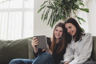 Two teenager sister girls are sitting at green sofa in living room and watching some funny video at