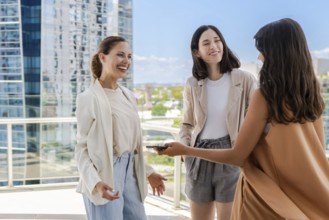 Three women are standing on a rooftop, smiling and talking to each other. One of them is holding a