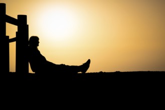 Silhouette of an unrecognizable person sitting against wooden pole with orange colored sunset in