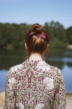 rear view of anonymous young woman with floral dress and red hair bun standing by lake in solitude