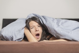 Tired young woman yawning under a duvet as she spends a lazy day relaxing at home