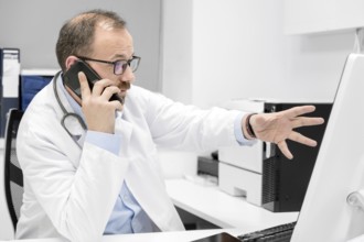 Male doctor pointing with finger on desktop computer while talking on the phone, discussing