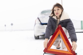 Woman puts up a warning triangle on a snowy road in winter, car breakdown in winter