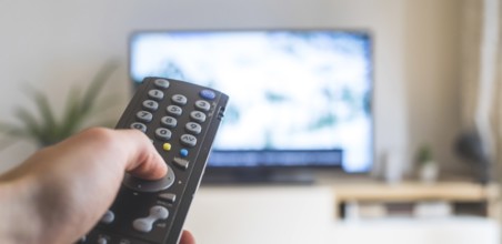 Holding a TV remote control in the hand, foreground, tv in the blurry background. Streaming