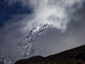 Two brightly lit snowy peaks peeking out of the clouds behind a dark ridge, Annapurna region,
