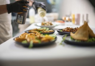 Chef wearing black gloves is pouring a sauce from a bottle on several plates of indian food,