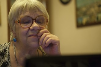Thoughtful senior woman with blond hair wearing spectacles reading a computer screen with her hand