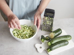 Woman with her hands in a bowl of grated zucchini, other fresh zucchini on wooden board and