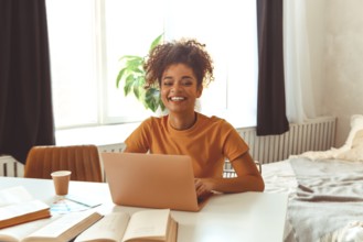 Cheerful young African American female student sitting at desk at home surrounded by textbooks, in