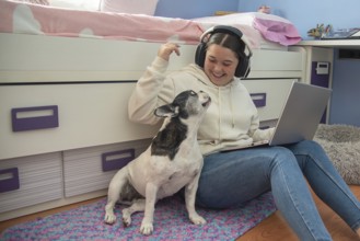 A teenager enjoys a moment of leisure in her room, working on her laptop and playing with her dog,