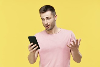 Shocked funny millennial man using mobile phone holding it in hand isolated over yellow background