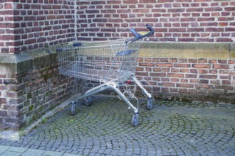 Metal shopping cart stands empty next to a brick wall on a cobblestone street, symbolizing