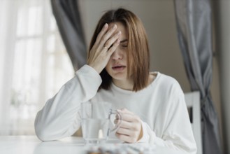 Woman holding her forehead, looking unwell, with a glass of water and curtains in the background