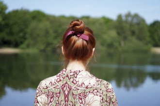 back view of unrecognizable young woman with floral dress and red hair bun looking at lake in