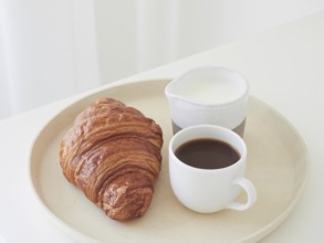Simple breakfast on a tray on the white table. Croissant, coffee cup and milk jug