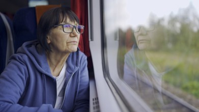 Elderly lady in glasses travels in train and looking out the window reflected in the glass