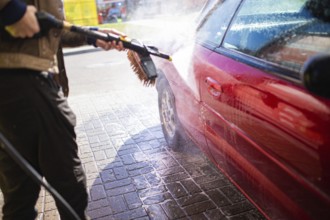 Pressure washing a red car on a cobblestone street in sunlight