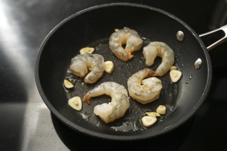 Raw peeled shrimps and garlic are sauteed in a cooking pan on the black stove, preparation for a