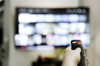 Hand holding remote control pointing to TV. Person watching smart television indoors. Blurred