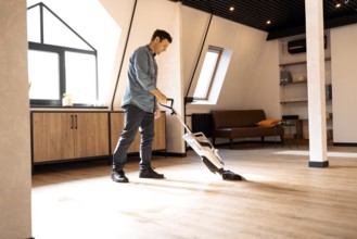 Brunette man vacuuming wooden floor in loft iving room with vacuum cleaner