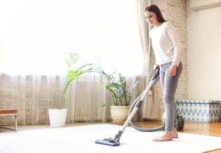 Full body young female using vacuum cleaner to tidy carpet in light room at home