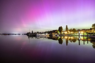 Colourful northern lights reflected in a quiet night lake next to illuminated buildings, harbour,