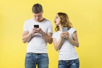 Suspicious girlfriend spying his boyfriend texting mobile phone against yellow backdrop
