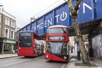 Red double decker buses driving on a london street, passing under a bridge with the camden town